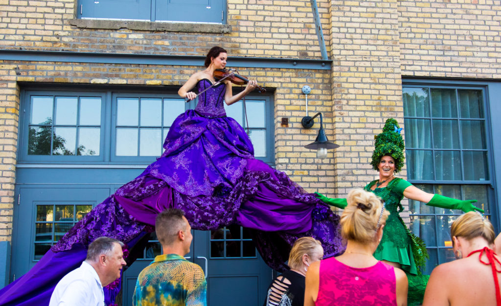 Purple violinist archway installation Enticing Entertainment provided for their event showcase. This event was held an the Machine Shop venue in Minneapolis. These characters greeted guests as they entered underneath the dress of the violinist.